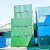 A stack of colorful shipping containers, primarily green and blue, against a clear sky and building backdrop.