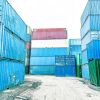 A container yard filled with stacked blue, green, and red shipping containers under a bright sky.
